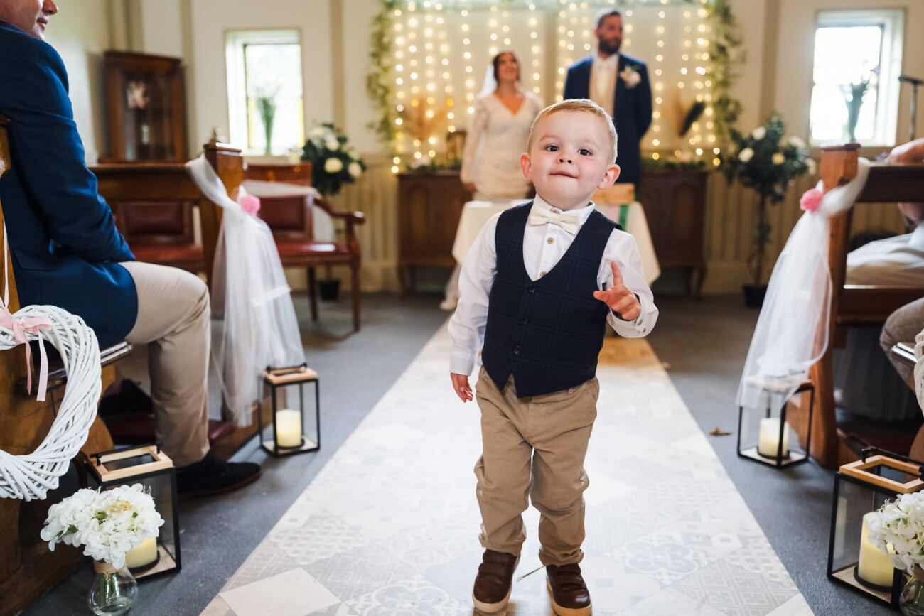 A Relaxed Summer Wedding at Village Barn, Tyrellspass | Amy & Eoghan 44 A young boy in formal attire walks down the aisle at a wedding ceremony, with a bride and groom standing in the background.