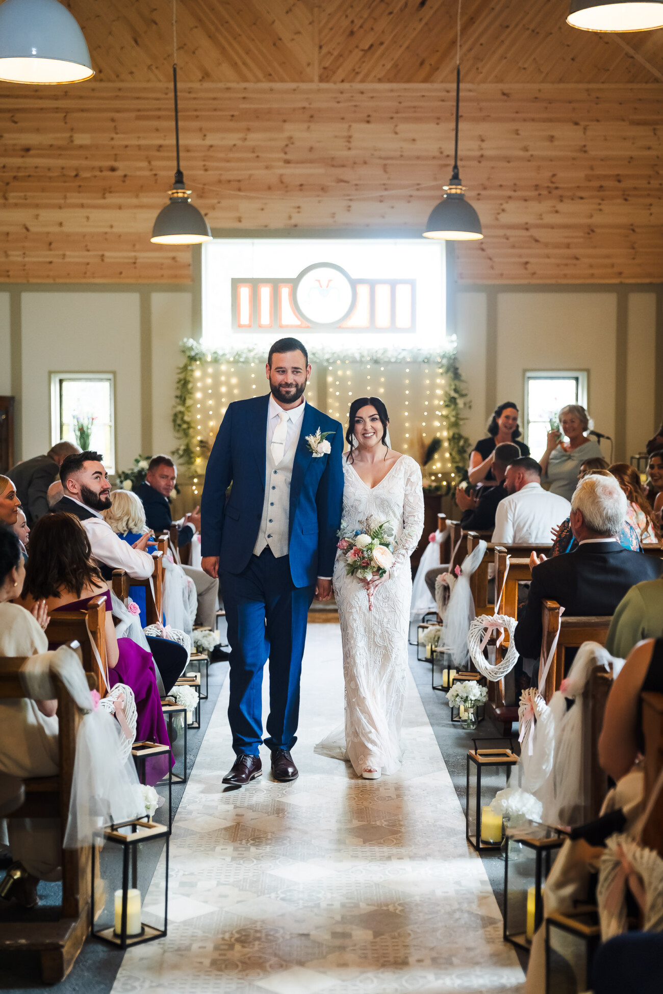 A Relaxed Summer Wedding at Village Barn, Tyrellspass | Amy & Eoghan 45 A bride and groom walk down the aisle together in a decorated wooden chapel, smiling at guests seated on both sides.