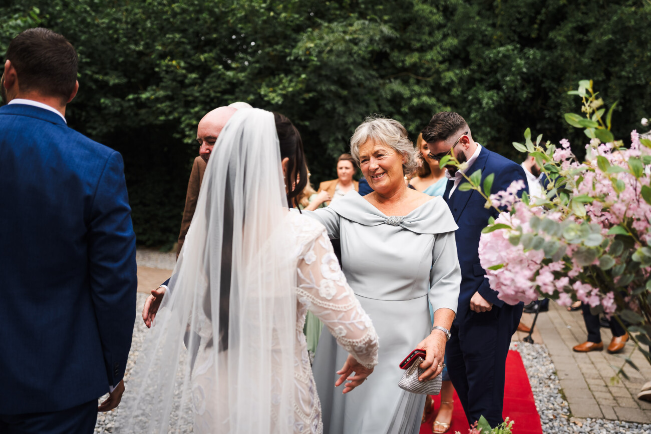 A Relaxed Summer Wedding at Village Barn, Tyrellspass | Amy & Eoghan 46 A bride in a white dress and veil greets an older woman in a light grey gown outdoors, with guests standing nearby and pink flowers in the foreground.
