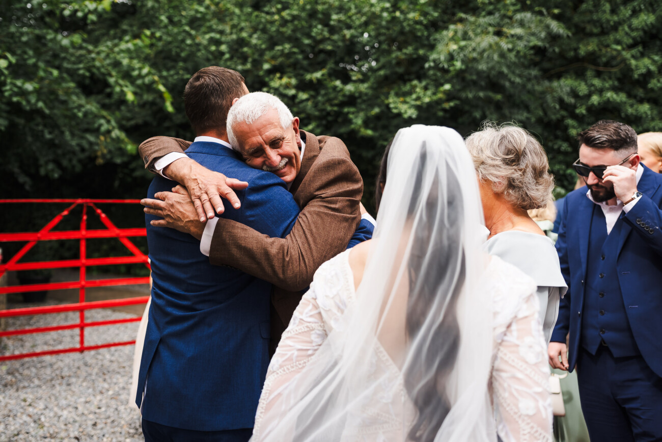A Relaxed Summer Wedding at Village Barn, Tyrellspass | Amy & Eoghan 47 An elderly man hugs a man in a blue suit at an outdoor event, while a bride in a white dress and veil stands nearby. Other guests are also present.