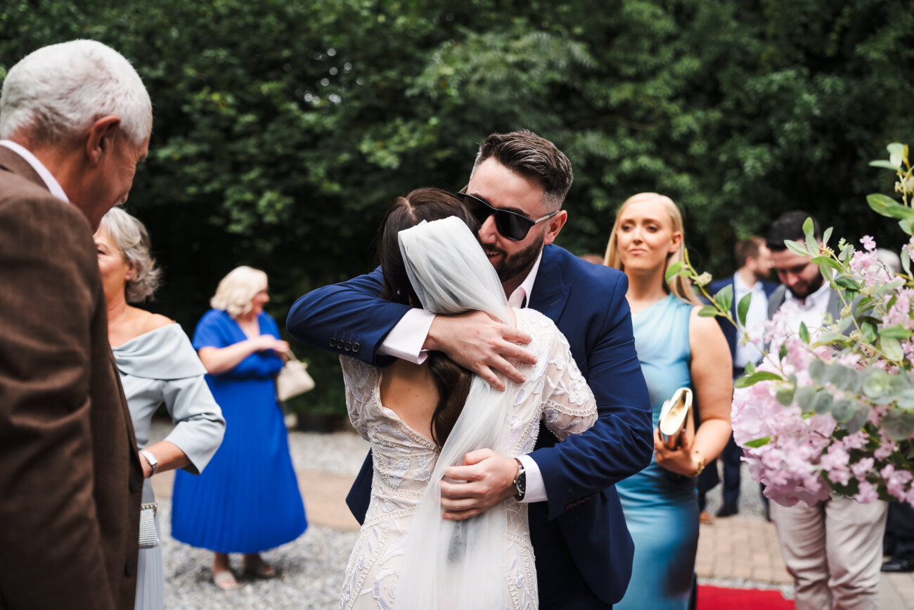 A Relaxed Summer Wedding at Village Barn, Tyrellspass | Amy & Eoghan 48 A man in sunglasses hugs a bride in a white dress and veil outdoors, with guests in formal attire standing nearby.