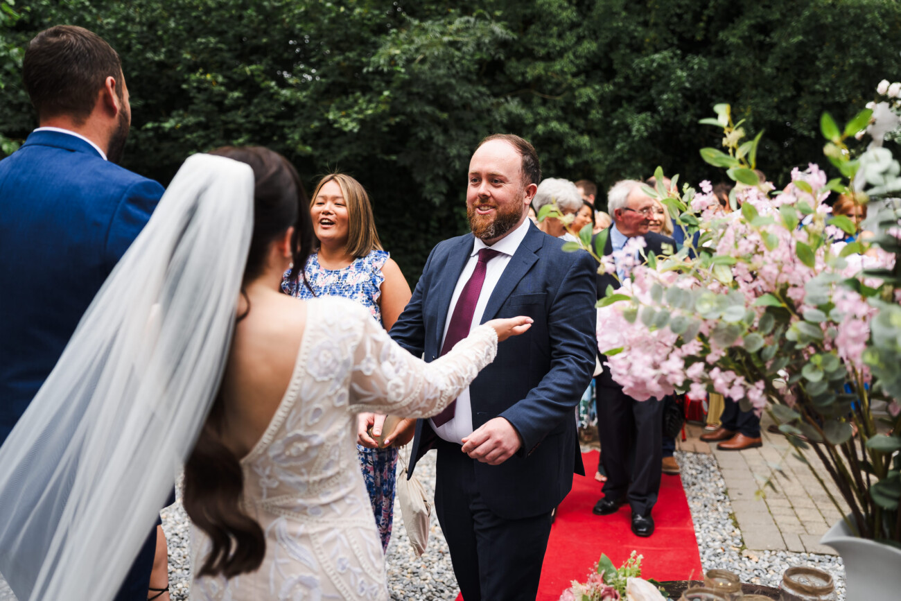 A Relaxed Summer Wedding at Village Barn, Tyrellspass | Amy & Eoghan 49 A bride in a white dress and veil greets a man in a suit on a red carpet outdoors, with other guests and flowers visible in the background.