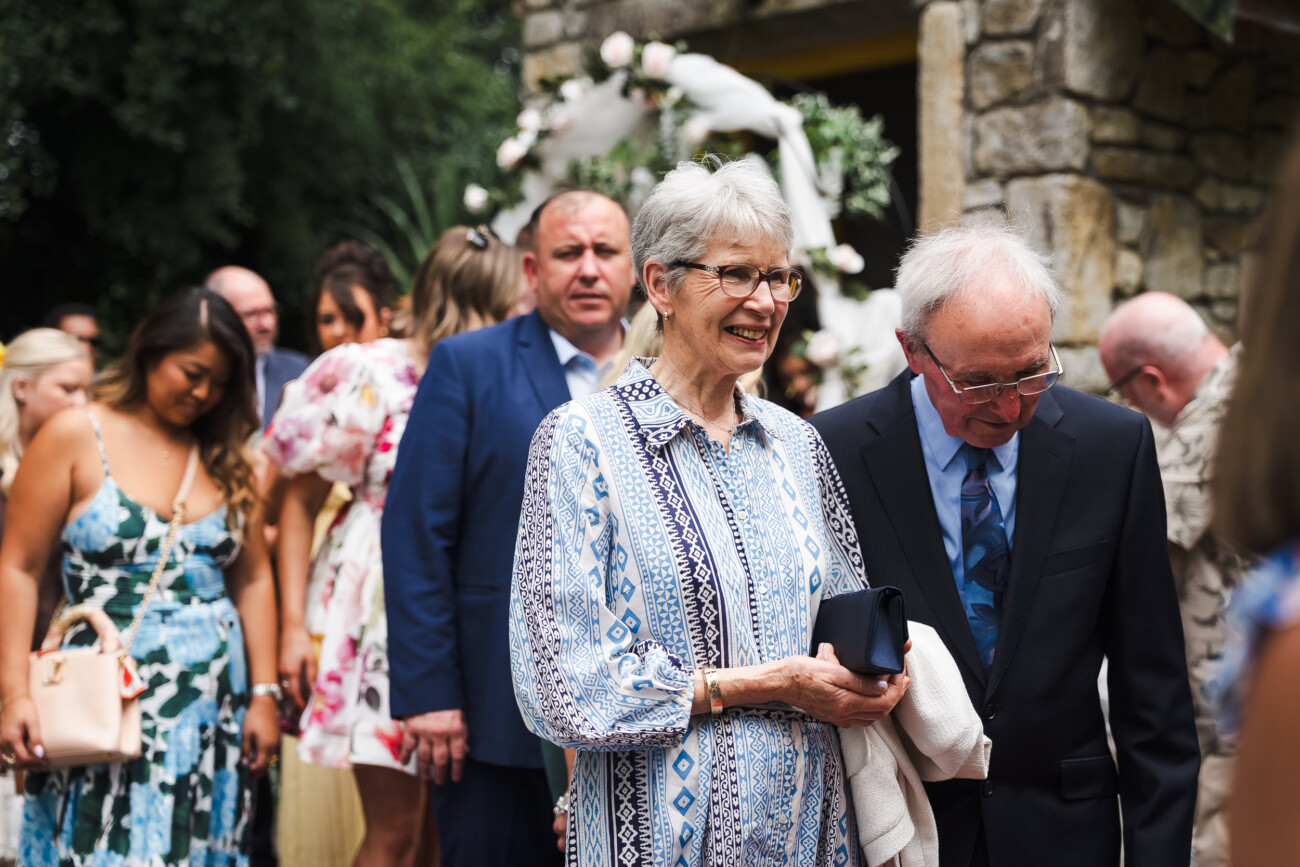 A Relaxed Summer Wedding at Village Barn, Tyrellspass | Amy & Eoghan 50 Older woman and man stand smiling at an outdoor event, with a crowd of people and a floral arch in the background.