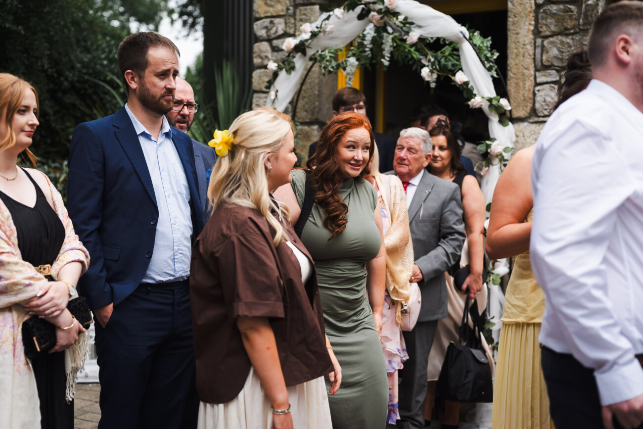 A Relaxed Summer Wedding at Village Barn, Tyrellspass | Amy & Eoghan 51 A group of people, dressed in semi-formal attire, stand outside near a stone building with a floral arch, appearing to queue or gather for an event.