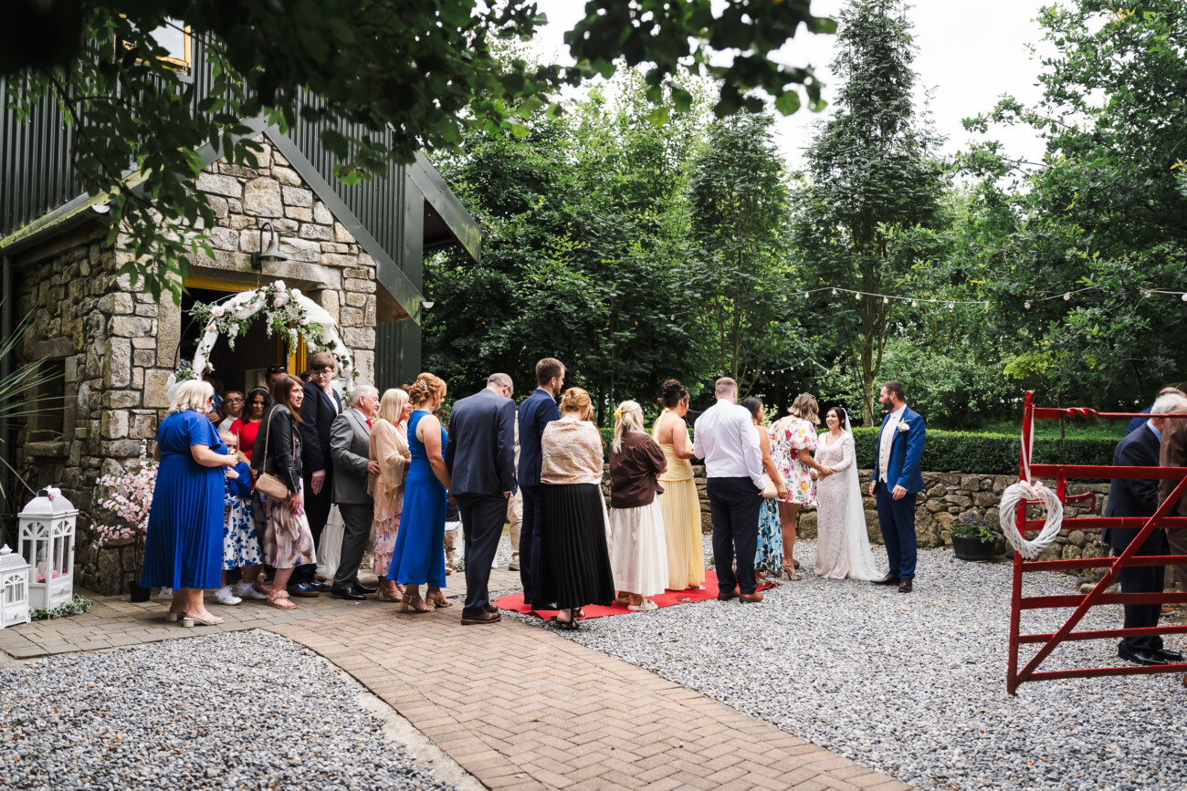 A Relaxed Summer Wedding at Village Barn, Tyrellspass | Amy & Eoghan 52 Guests queue outside a stone building decorated with flowers, waiting to greet a bride and groom at a wedding reception.