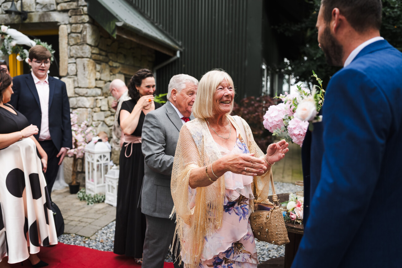 A Relaxed Summer Wedding at Village Barn, Tyrellspass | Amy & Eoghan 53 An older woman in a beige shawl smiles and extends her hands to greet a man in a blue suit at an outdoor event with other guests nearby.