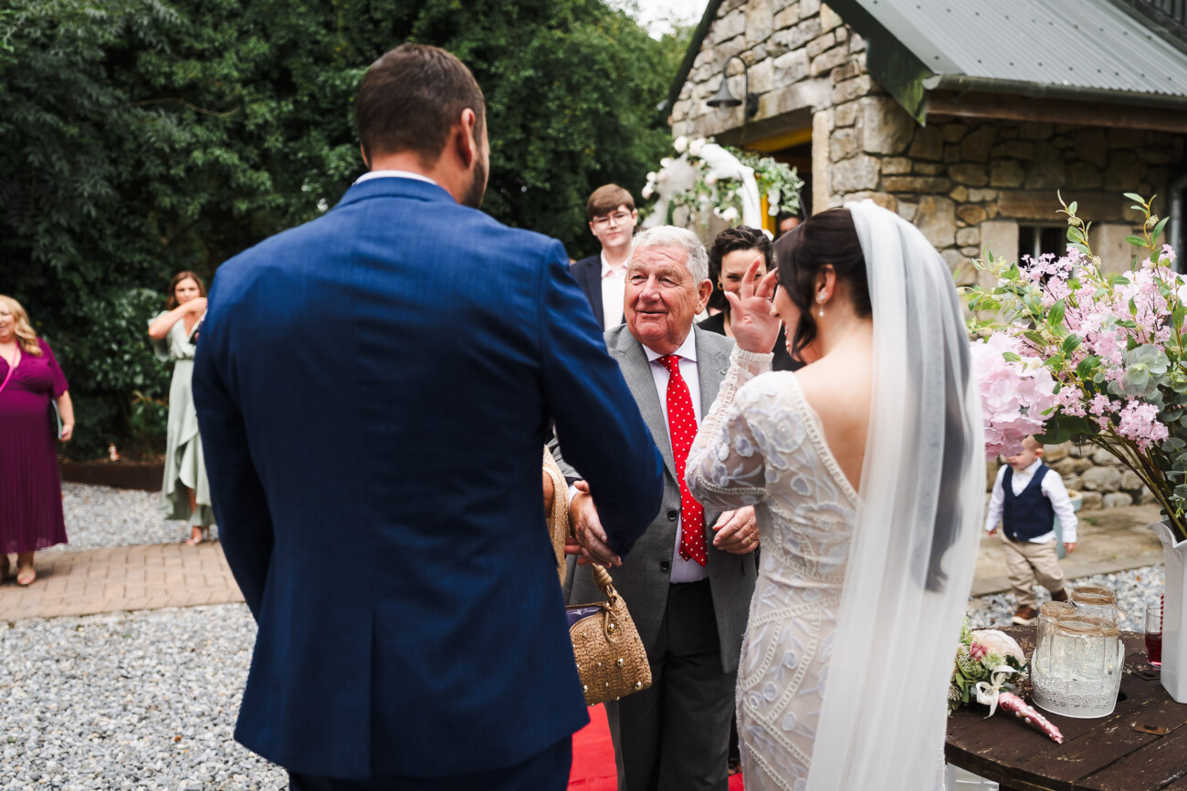 A Relaxed Summer Wedding at Village Barn, Tyrellspass | Amy & Eoghan 54 A bride in a white dress and veil greets an older man in a suit and red spotted tie outdoors, with guests and a stone building in the background.