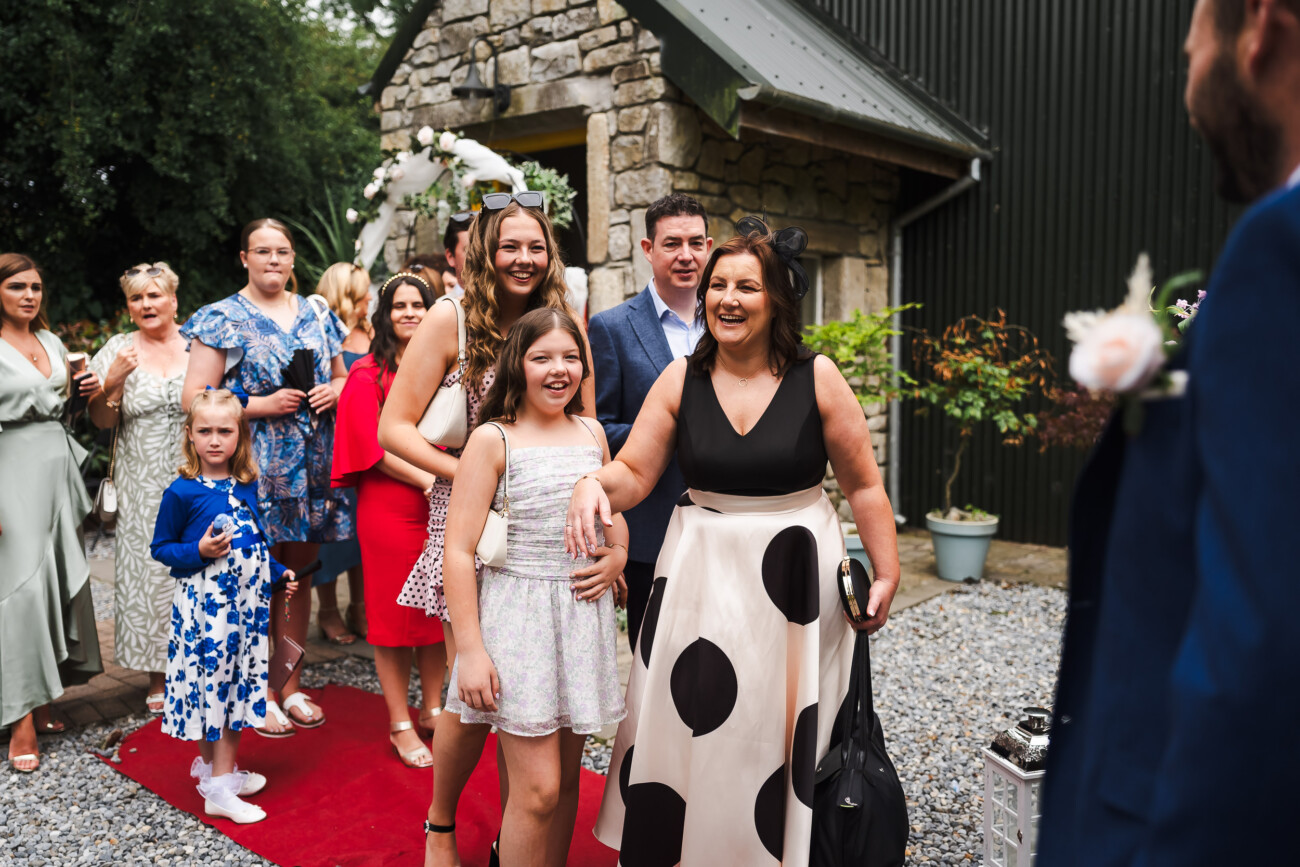 A Relaxed Summer Wedding at Village Barn, Tyrellspass | Amy & Eoghan 55 A group of people, including women and children, stand in a queue on a red carpet outside a stone building, smiling and waiting for their turn to greet someone dressed in a blue suit.