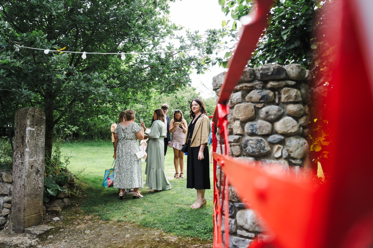 A Relaxed Summer Wedding at Village Barn, Tyrellspass | Amy & Eoghan 56 A group of people stand and talk on a grassy lawn near a stone gate and red ribbon, surrounded by trees and greenery.