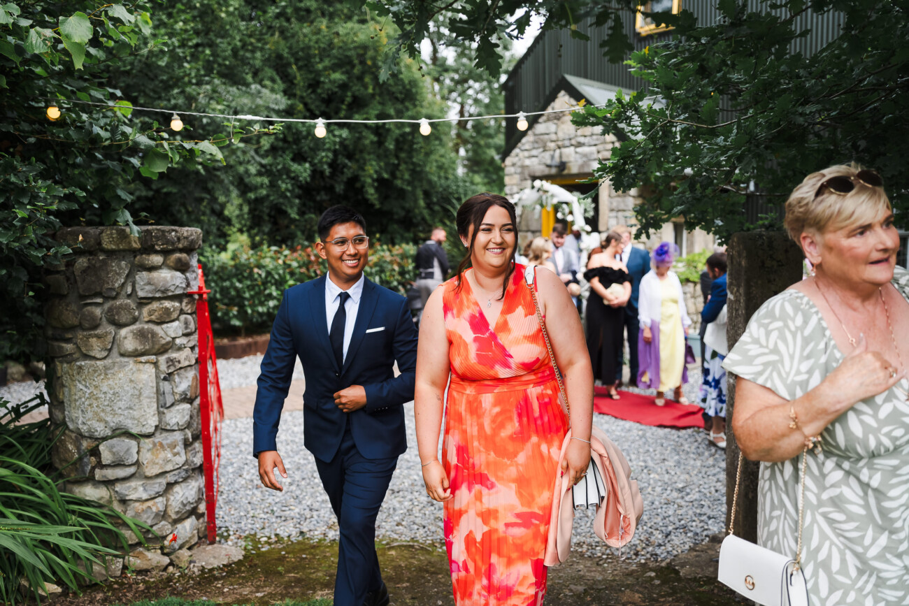 A Relaxed Summer Wedding at Village Barn, Tyrellspass | Amy & Eoghan 57 A man in a suit and a woman in a red floral dress walk on a stone path at an outdoor event, with other guests and fairy lights in the background.