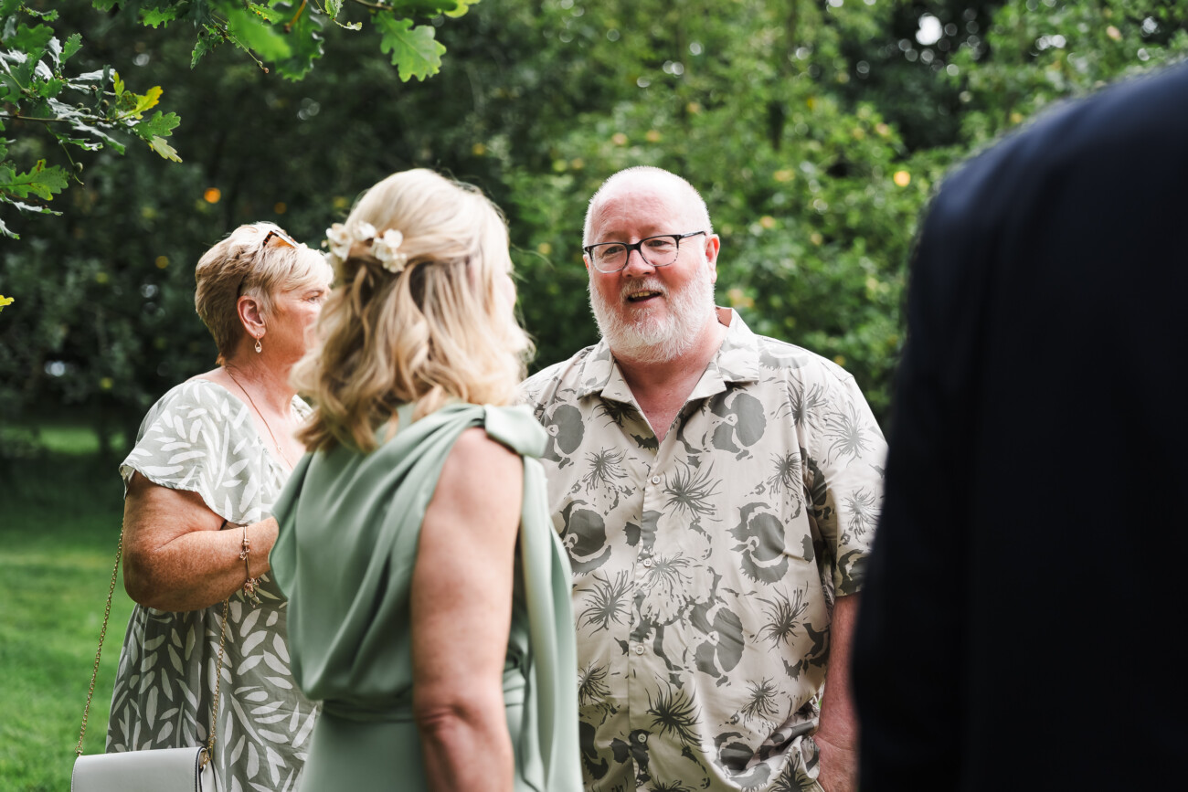 A Relaxed Summer Wedding at Village Barn, Tyrellspass | Amy & Eoghan 58 A man in a patterned shirt talks with two women outdoors; trees and greenery are in the background.