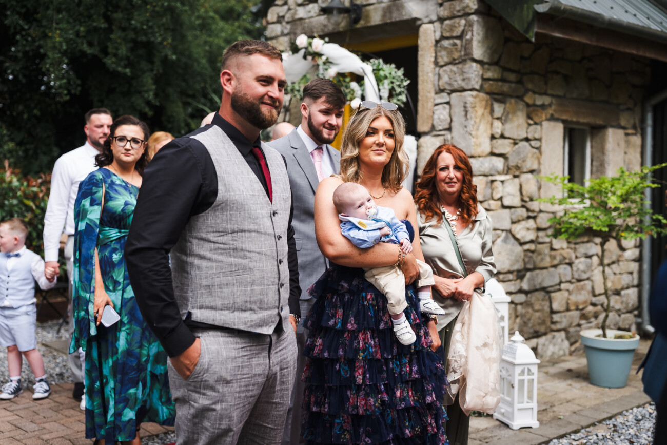 A Relaxed Summer Wedding at Village Barn, Tyrellspass | Amy & Eoghan 59 A group of well-dressed adults and a baby stand outdoors by a stone building, posing for a photo during a daytime event.