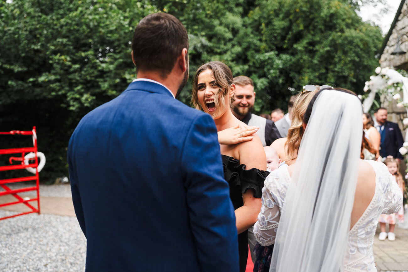 A Relaxed Summer Wedding at Village Barn, Tyrellspass | Amy & Eoghan 60 A woman in a black dress appears surprised whilst talking to a man in a blue suit and a bride in a white dress outdoors, with other guests and greenery in the background.