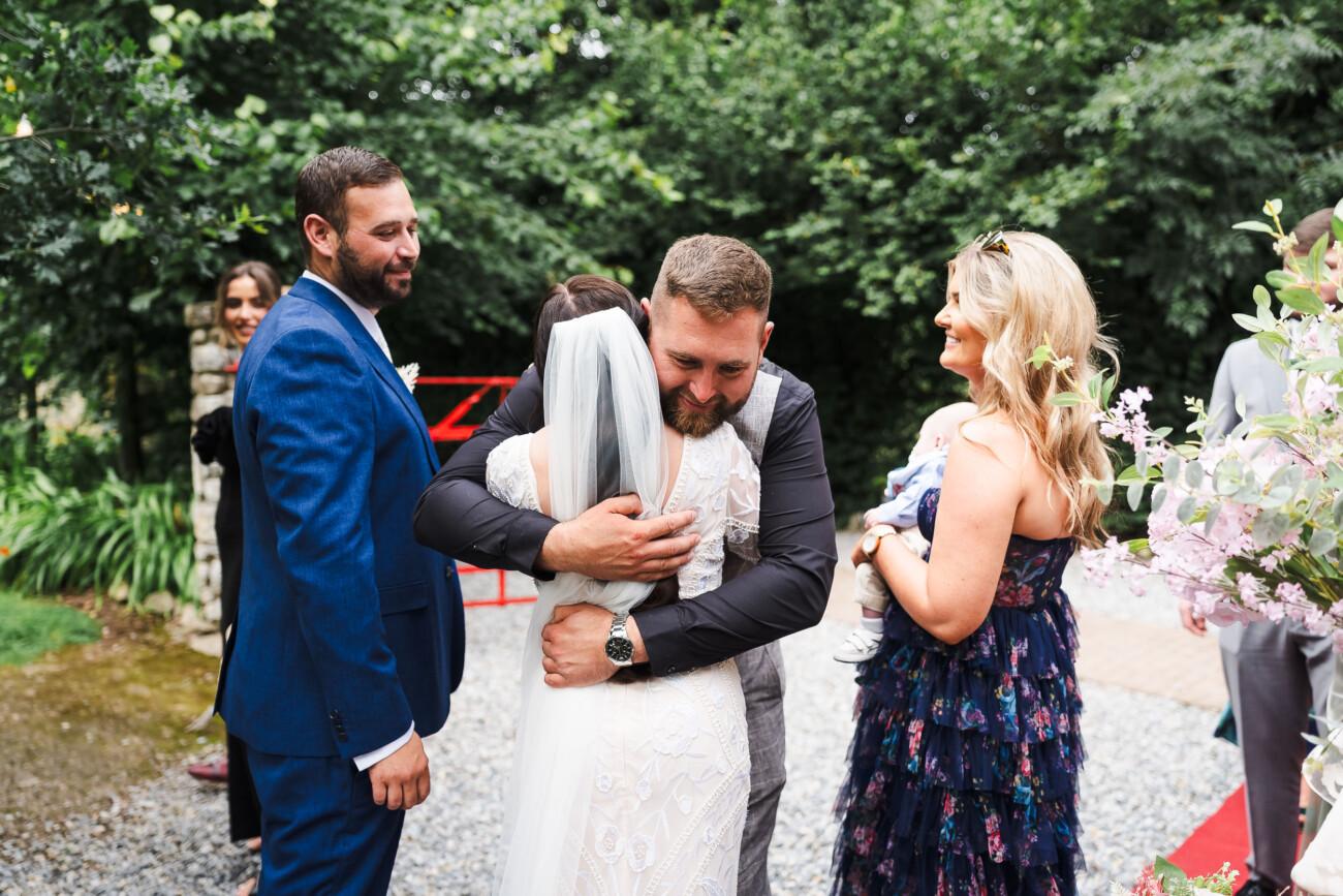 A Relaxed Summer Wedding at Village Barn, Tyrellspass | Amy & Eoghan 61 A man hugs a bride outdoors as guests in formal attire stand nearby, smiling amid greenery and flowers.