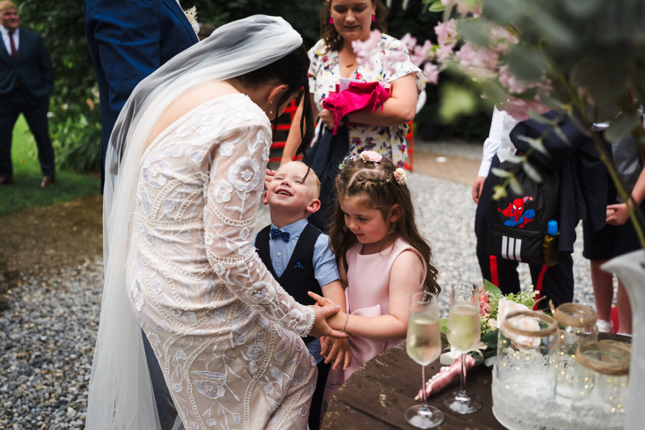 A Relaxed Summer Wedding at Village Barn, Tyrellspass | Amy & Eoghan 62 A bride in a white dress bends down to greet a smiling young boy and girl at an outdoor gathering, surrounded by guests and champagne on a table.