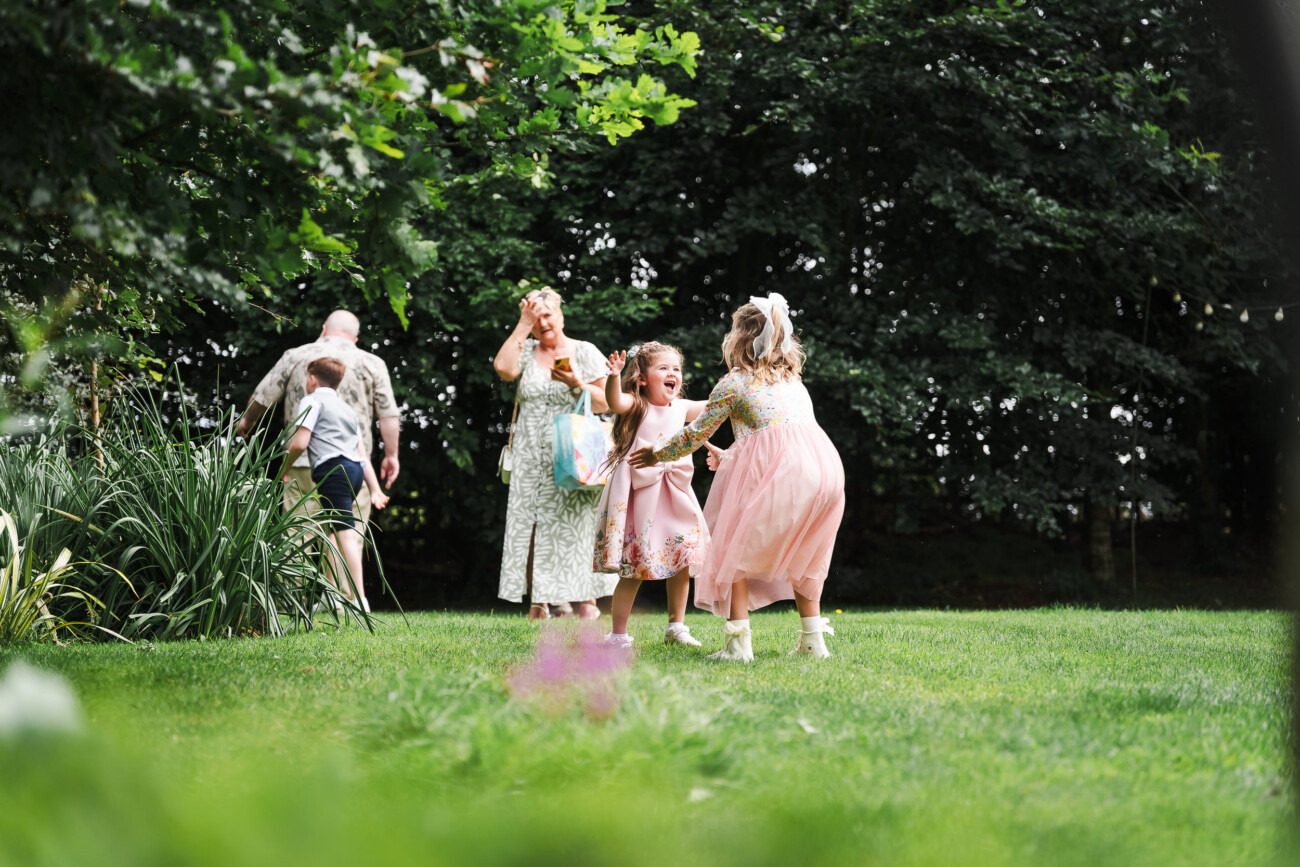 A Relaxed Summer Wedding at Village Barn, Tyrellspass | Amy & Eoghan 63 Two girls in dresses play and laugh on a grassy lawn, while two adults and another child walk in the background near trees and plants.