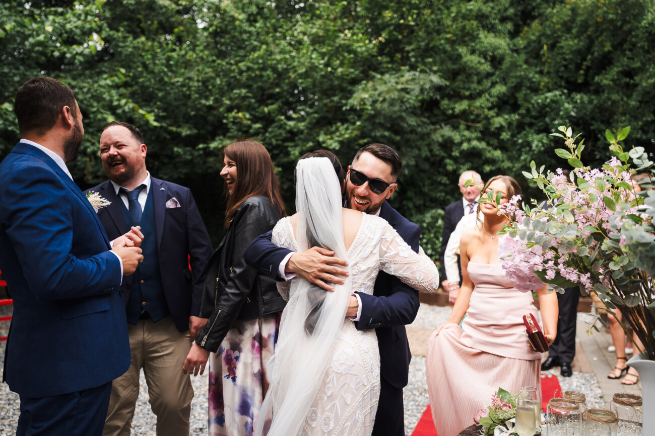 A Relaxed Summer Wedding at Village Barn, Tyrellspass | Amy & Eoghan 64 A bride in a white dress hugs a guest in sunglasses at an outdoor wedding, surrounded by other smiling guests near a table with floral decorations.