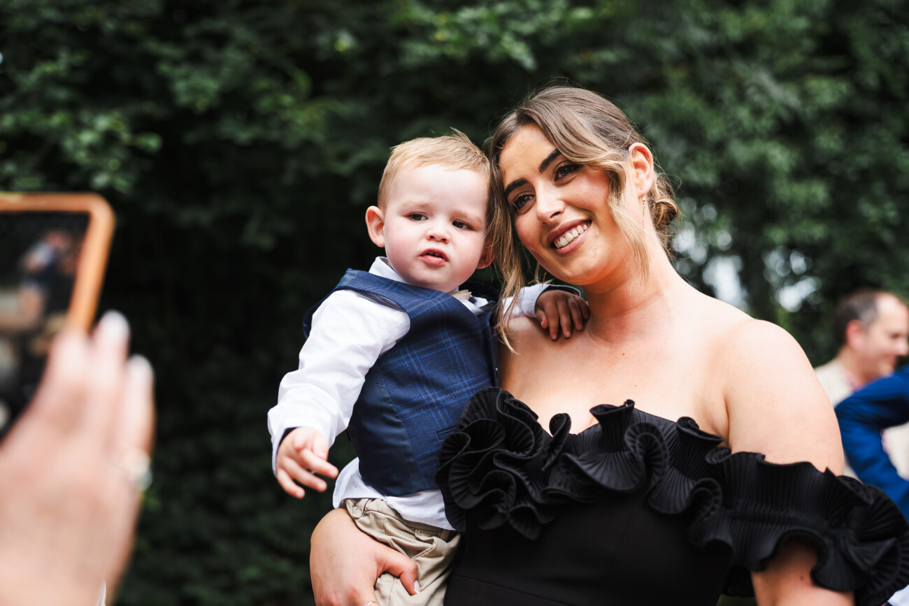 A Relaxed Summer Wedding at Village Barn, Tyrellspass | Amy & Eoghan 65 A woman in a black dress holds a young boy in smart clothes while smiling for a photo outdoors.