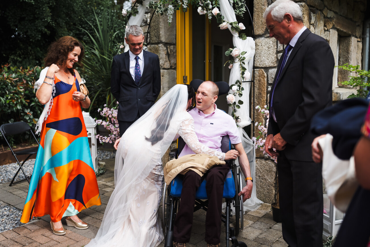 A Relaxed Summer Wedding at Village Barn, Tyrellspass | Amy & Eoghan 66 A bride in a white dress and veil bends to hug a man in a wheelchair outside, surrounded by three formally dressed guests.