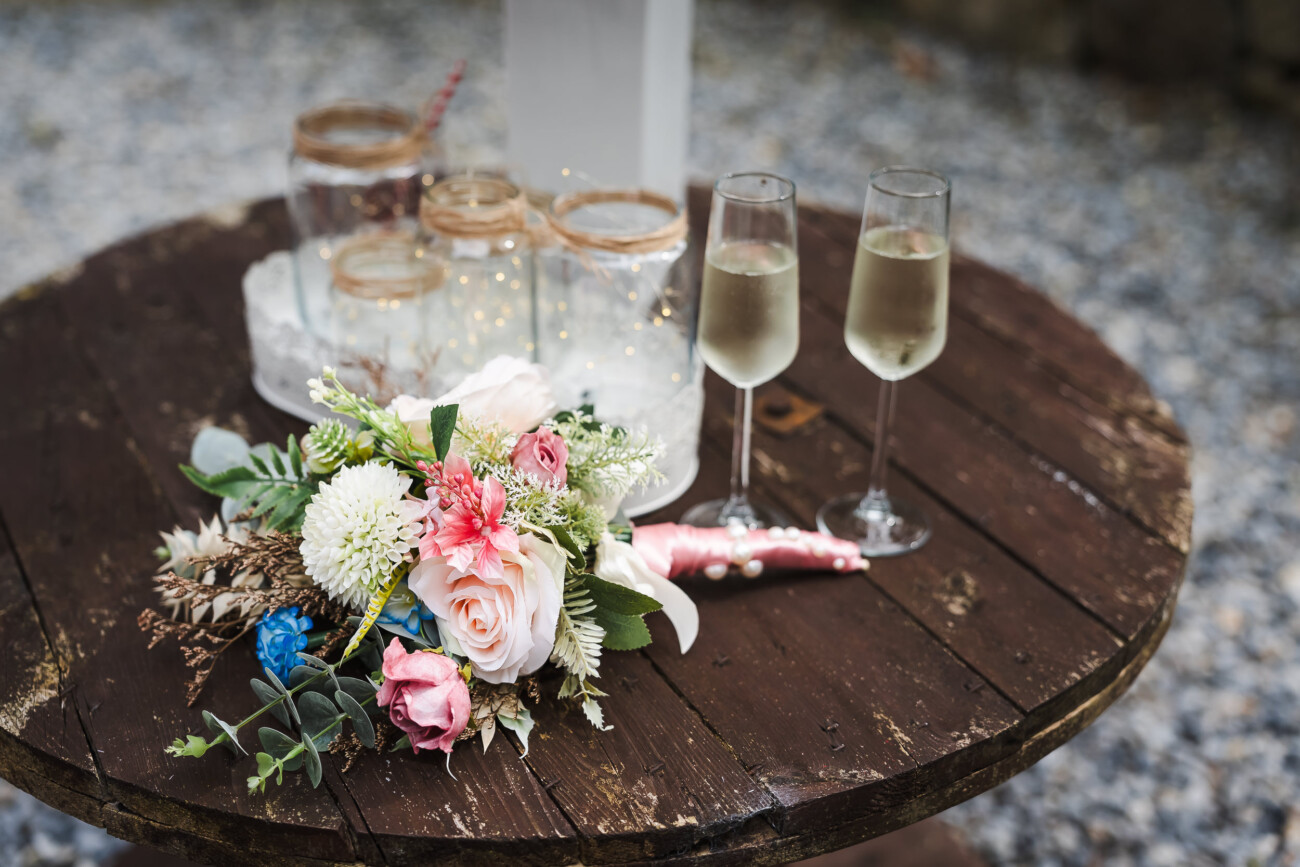 A Relaxed Summer Wedding at Village Barn, Tyrellspass | Amy & Eoghan 67 A bouquet, two glasses of champagne, and jars with string sit on a round wooden table with a gravel background.