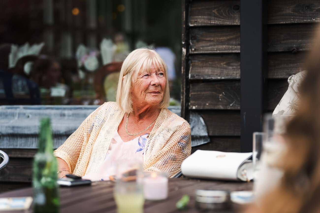 A Relaxed Summer Wedding at Village Barn, Tyrellspass | Amy & Eoghan 79 Elderly woman with blonde hair sits at an outdoor wooden table with drinks and a mobile, wearing a light cardigan and looking to the side.