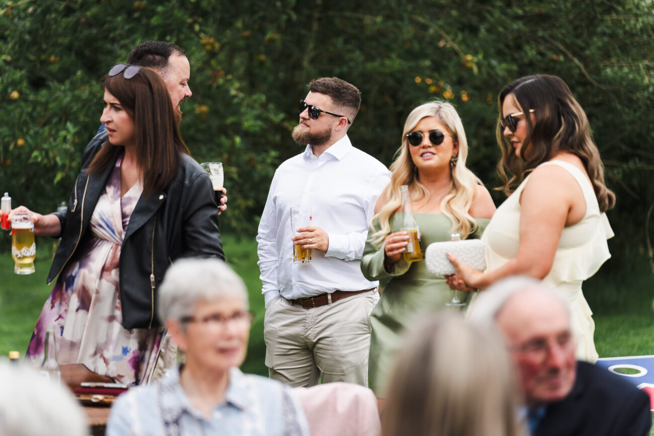 A Relaxed Summer Wedding at Village Barn, Tyrellspass | Amy & Eoghan 80 Five people stand outdoors, holding drinks and conversing, while others sit blurred in the foreground. The group is dressed in semi-formal attire on a grassy area with trees in the background.
