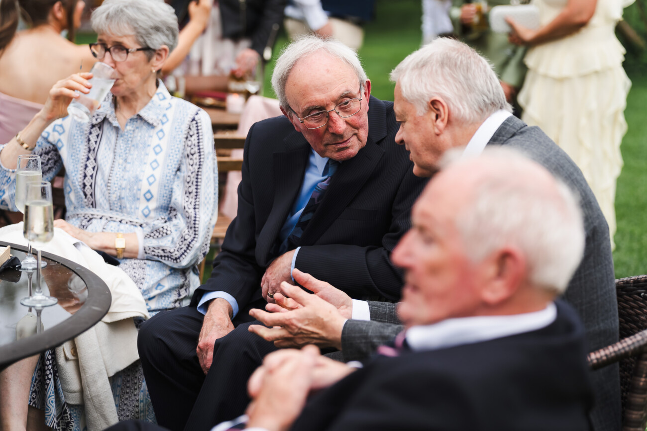 A Relaxed Summer Wedding at Village Barn, Tyrellspass | Amy & Eoghan 82 Four older adults sit outdoors at a table, engaged in conversation. One woman is drinking from a glass, whilst two men are talking closely. The setting appears to be a garden event.