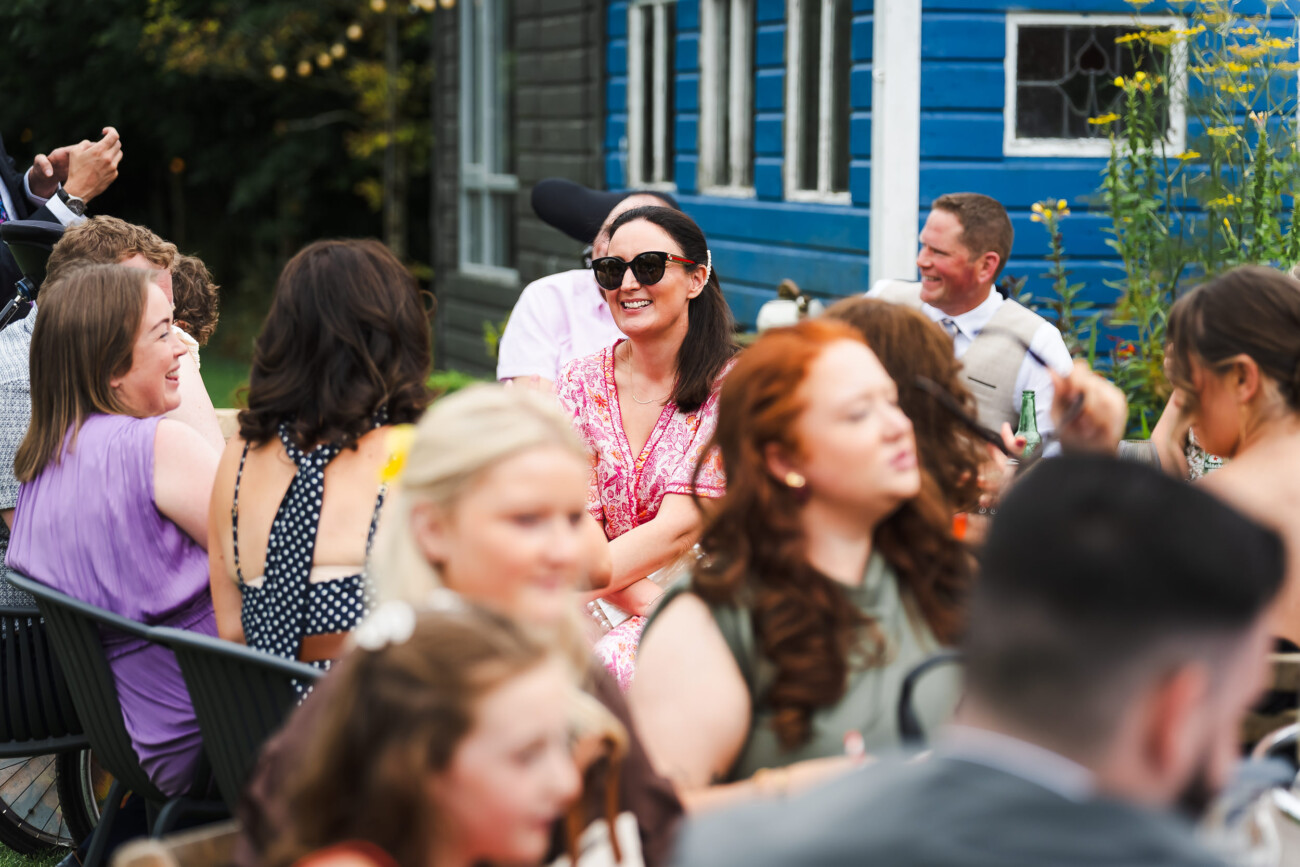 A Relaxed Summer Wedding at Village Barn, Tyrellspass | Amy & Eoghan 83 People sit and talk outdoors at a lively social gathering, with a woman in sunglasses and a pink dress smiling near the centre, and a blue building visible in the background.