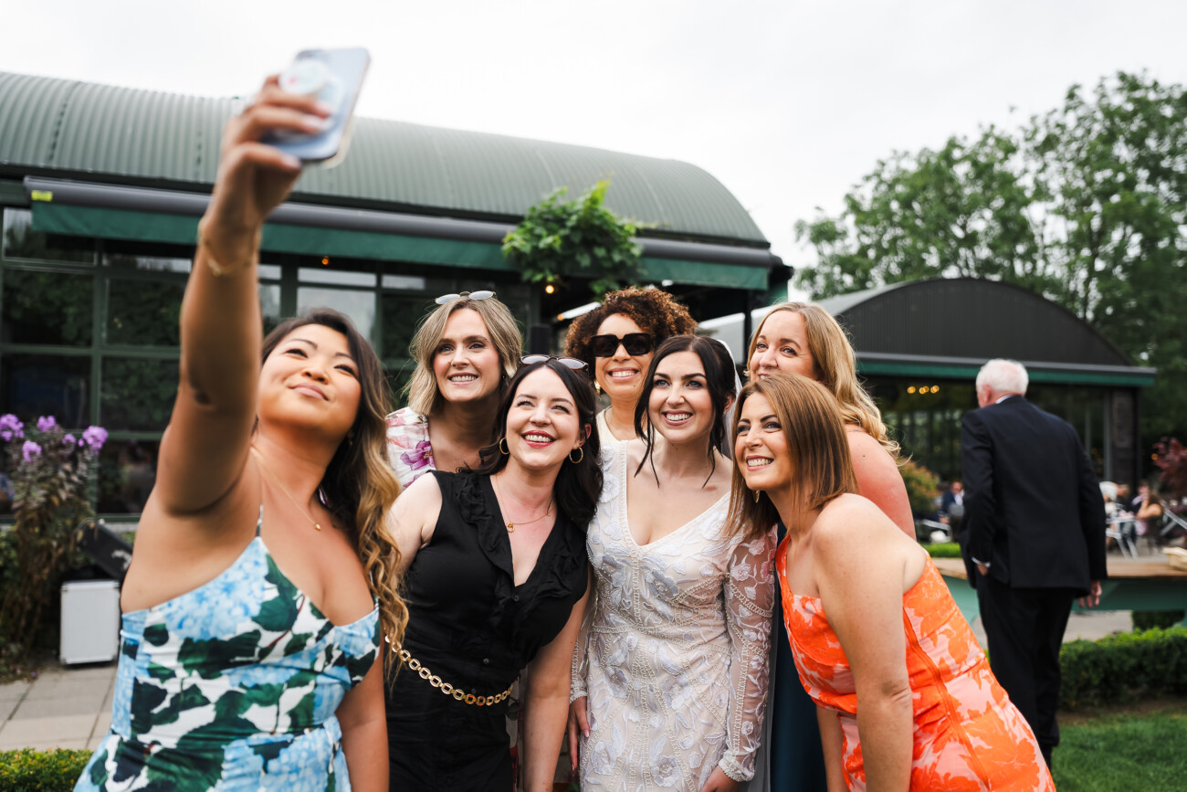 A Relaxed Summer Wedding at Village Barn, Tyrellspass | Amy & Eoghan 85 A group of women pose for a selfie outside at a social event, smiling at the camera with a building and greenery in the background.