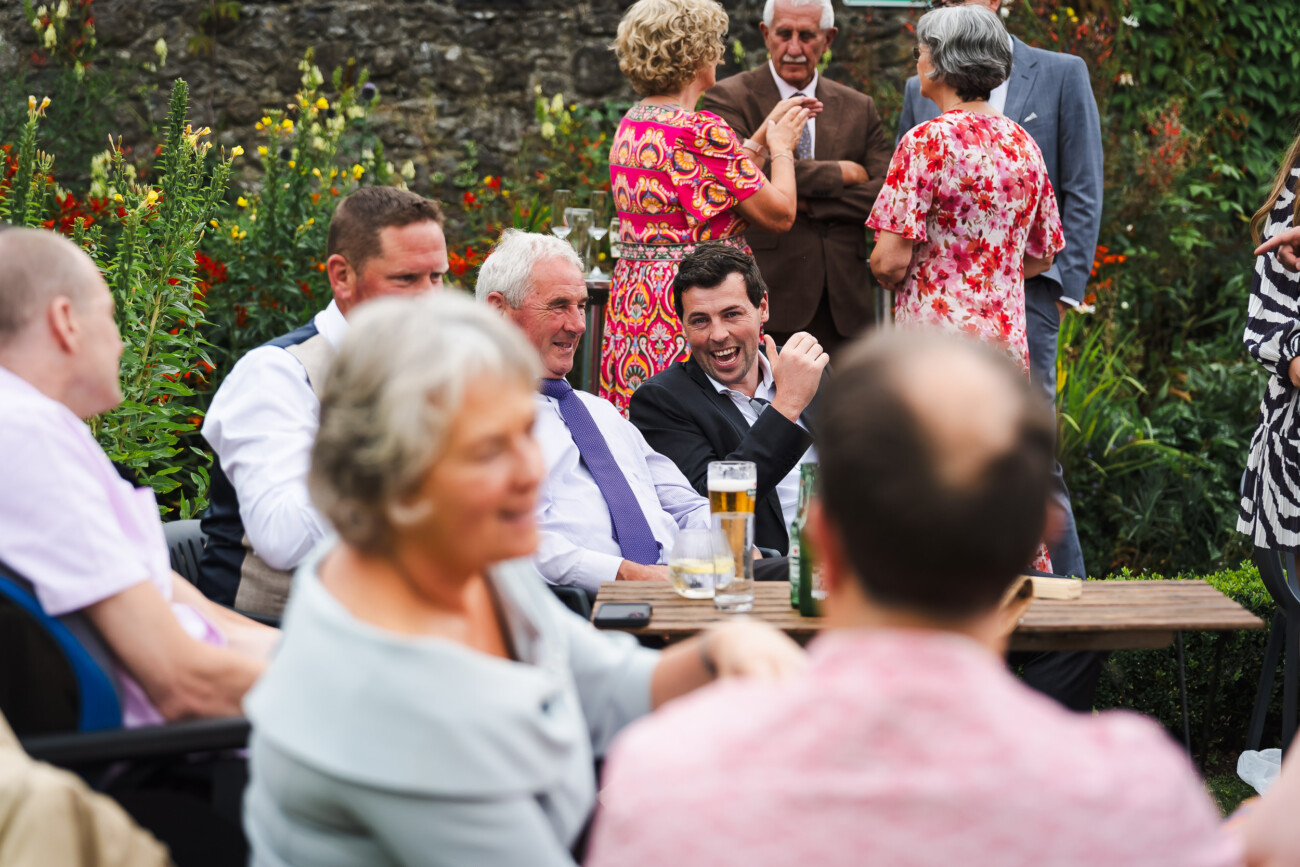 A Relaxed Summer Wedding at Village Barn, Tyrellspass | Amy & Eoghan 86 A group of people, both seated and standing, converse and laugh outdoors near a garden, with drinks on a table and greenery in the background.