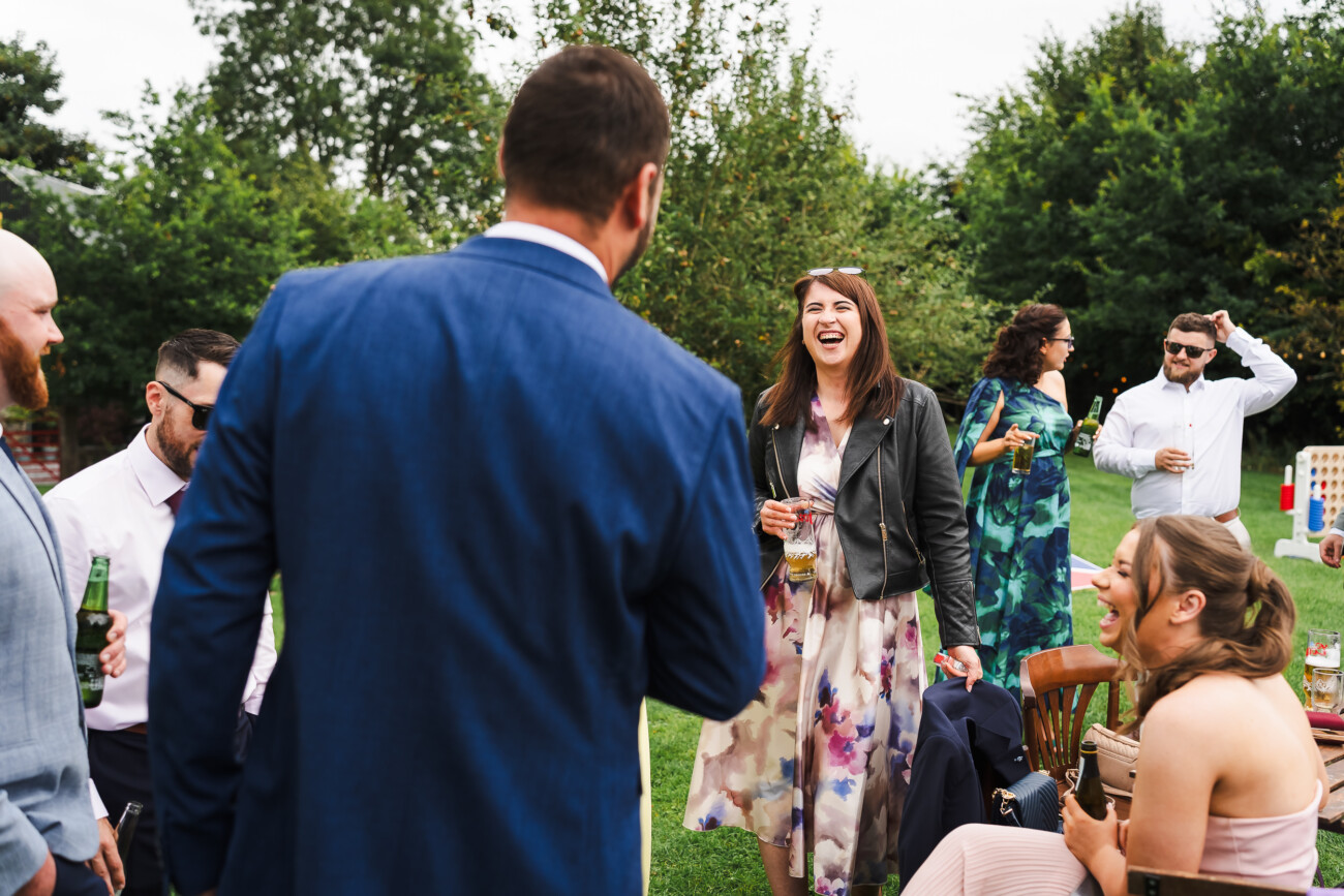 A Relaxed Summer Wedding at Village Barn, Tyrellspass | Amy & Eoghan 87 A group of people dressed in semi-formal attire socialise and laugh outdoors on a lawn, surrounded by trees and greenery, with drinks in hand.