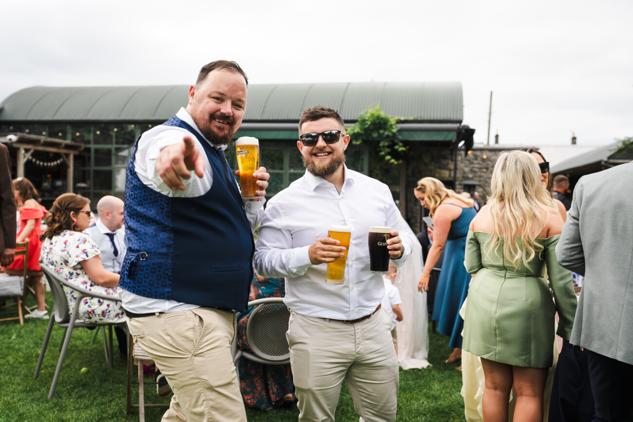 A Relaxed Summer Wedding at Village Barn, Tyrellspass | Amy & Eoghan 88 Two men stand on grass at an outdoor event, holding drinks and smiling at the camera; other guests in formal attire converse in the background.
