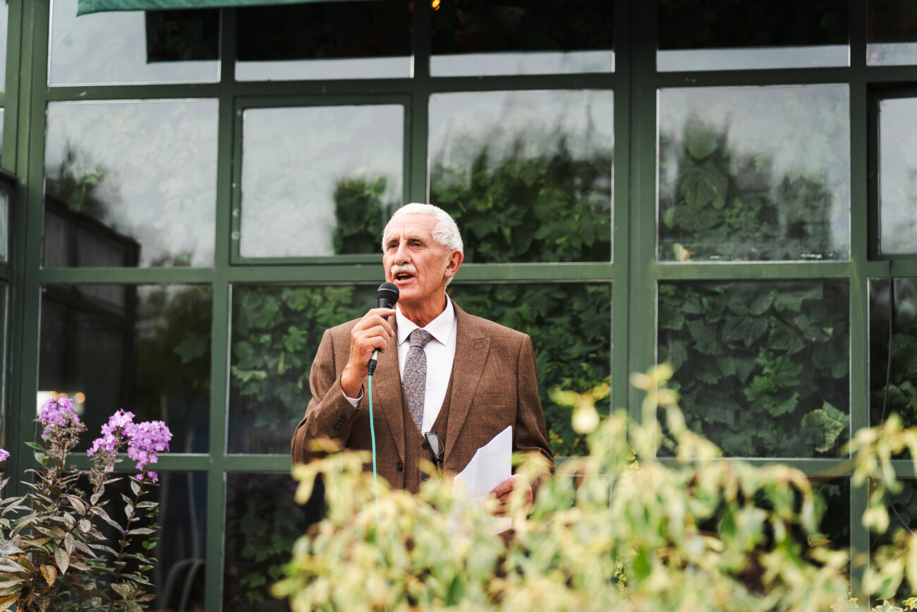 A Relaxed Summer Wedding at Village Barn, Tyrellspass | Amy & Eoghan 89 An older man in a brown suit speaks into a microphone whilst standing outdoors in front of a window with green foliage.
