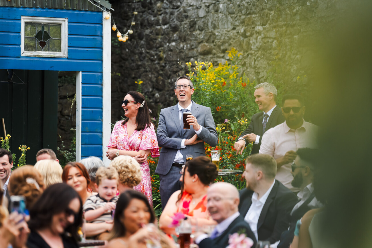 A Relaxed Summer Wedding at Village Barn, Tyrellspass | Amy & Eoghan 90 A group of people, some seated and some standing, socialise outdoors at a daytime event. A man in a suit laughs whilst holding a drink, and colourful flowers are visible in the background.