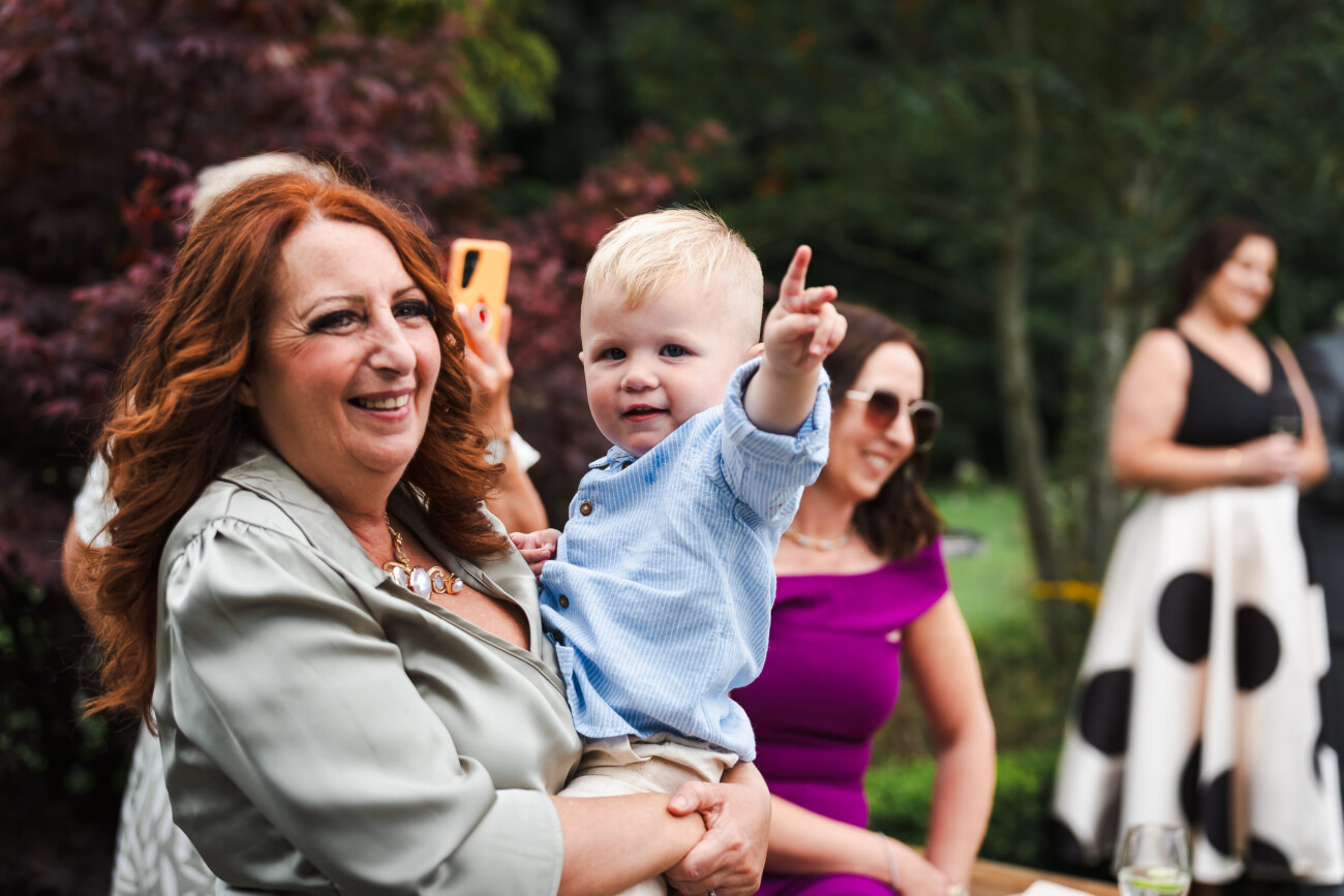 A Relaxed Summer Wedding at Village Barn, Tyrellspass | Amy & Eoghan 91 A woman with long red hair holds a smiling toddler who is pointing, with two women sitting in the background at an outdoor gathering.