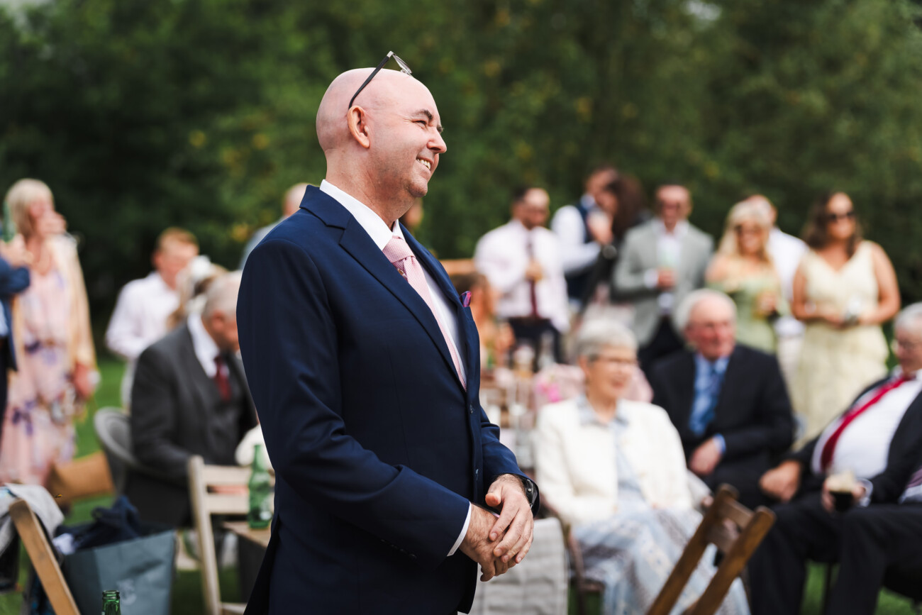 A Relaxed Summer Wedding at Village Barn, Tyrellspass | Amy & Eoghan 92 Bald man in a navy suit stands and smiles at an outdoor event with seated and standing guests visible in the background.