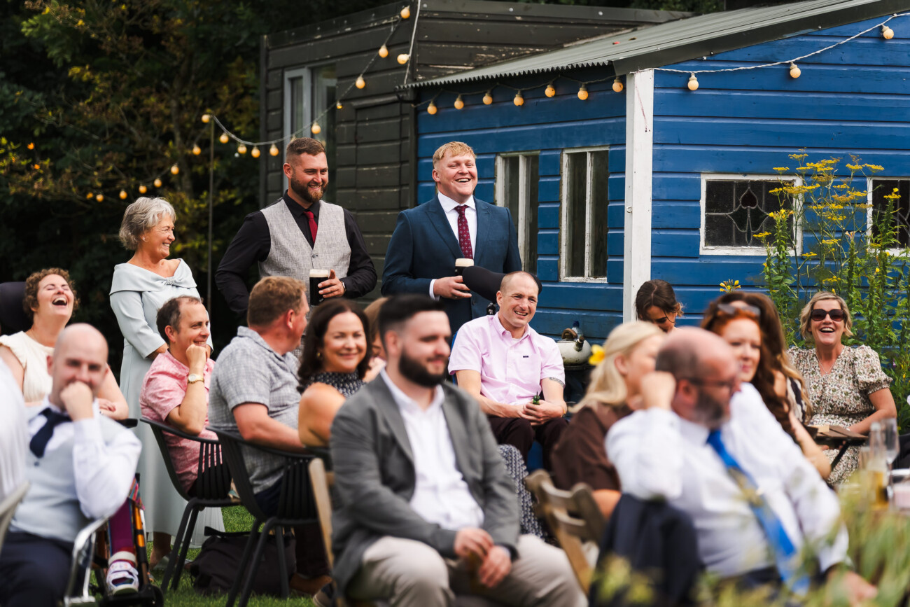 A Relaxed Summer Wedding at Village Barn, Tyrellspass | Amy & Eoghan 93 People seated outdoors at an event, laughing and smiling, with two men standing in the centre. A blue wooden building and fairy lights are visible in the background.