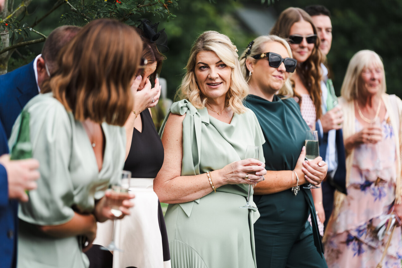 A Relaxed Summer Wedding at Village Barn, Tyrellspass | Amy & Eoghan 94 A group of people dressed in formal attire stand outdoors, holding drinks and conversing during a social event.