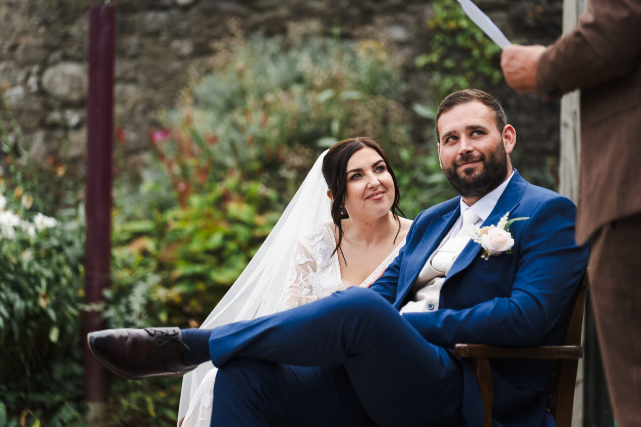 A Relaxed Summer Wedding at Village Barn, Tyrellspass | Amy & Eoghan 95 A bride and groom sit outdoors during a wedding ceremony, listening to a person speaking whilst surrounded by greenery and stone walls.