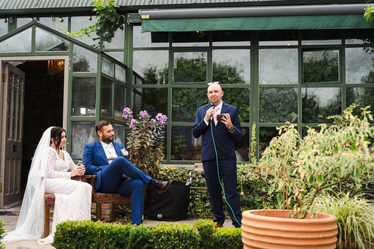 A Relaxed Summer Wedding at Village Barn, Tyrellspass | Amy & Eoghan 96 A man gives a speech outdoors with a microphone whilst a bride and groom sit nearby, listening in front of a glass-walled building with greenery.