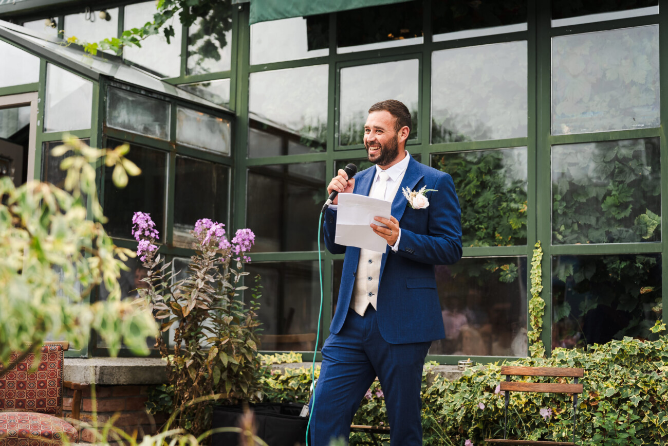 A Relaxed Summer Wedding at Village Barn, Tyrellspass | Amy & Eoghan 97 A man in a blue suit stands outside holding a microphone and papers, speaking in front of a glass building with plants and flowers nearby.