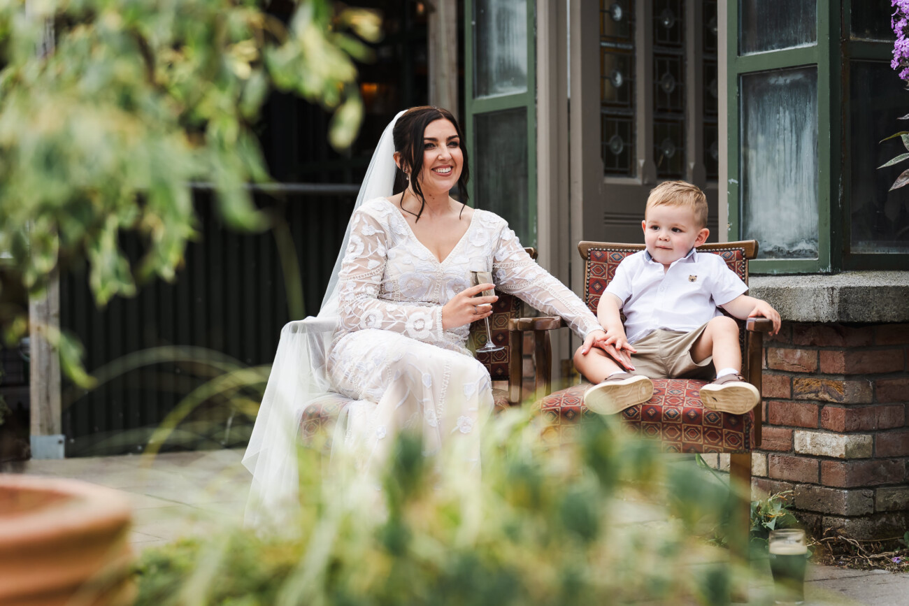 A Relaxed Summer Wedding at Village Barn, Tyrellspass | Amy & Eoghan 98 A bride in a white dress sits outdoors next to a young boy on a chair. She holds a glass and smiles, whilst the boy looks away. Greenery and a building are in the background.