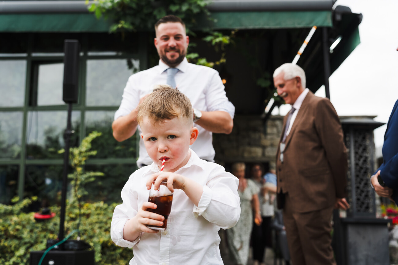 A Relaxed Summer Wedding at Village Barn, Tyrellspass | Amy & Eoghan 99 A young boy in a white shirt drinks from a straw, standing outdoors with adults in the background near a building.
