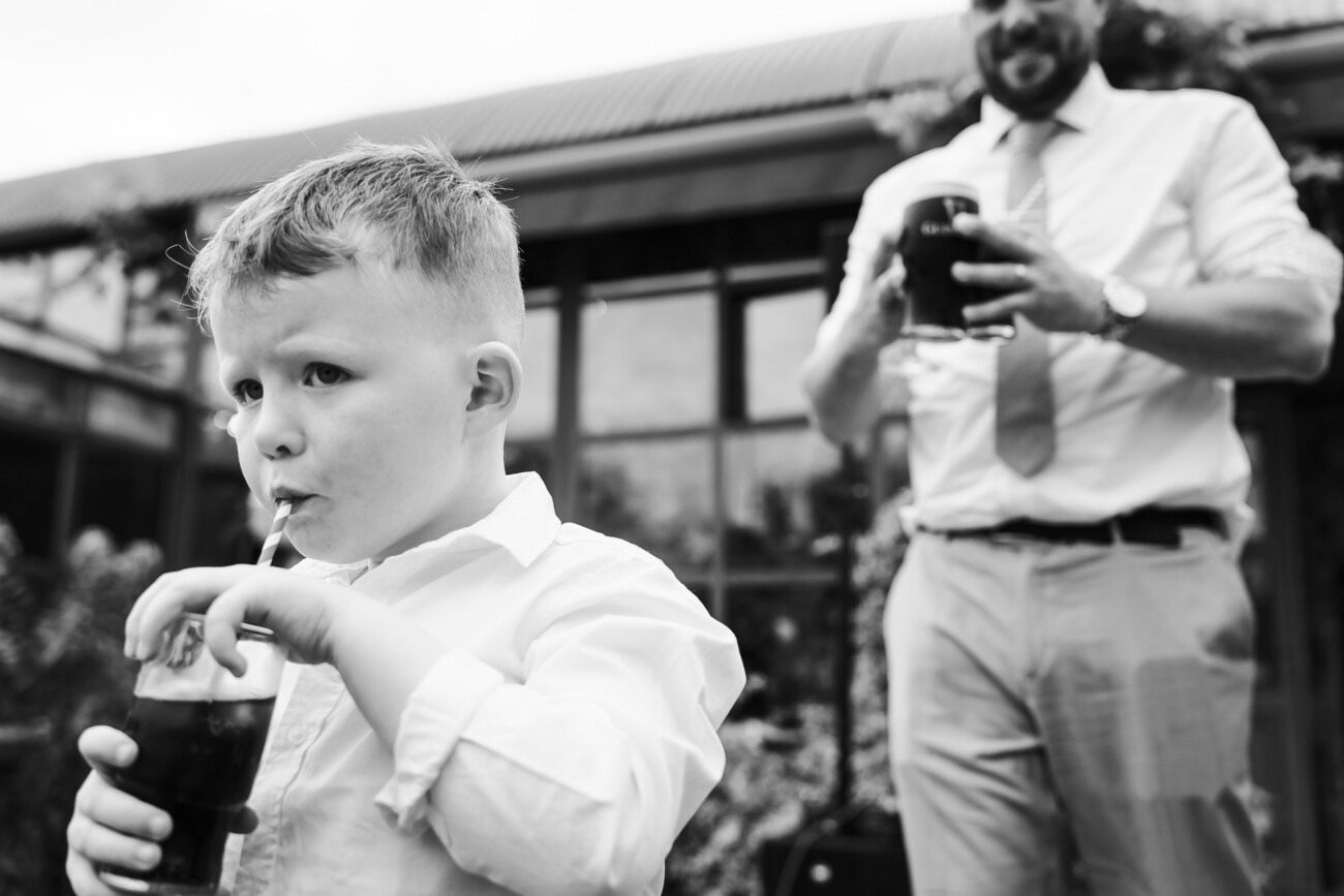 A Relaxed Summer Wedding at Village Barn, Tyrellspass | Amy & Eoghan 100 A young boy drinks from a glass with a straw in the foreground, whilst a man holding a glass stands in the blurred background.