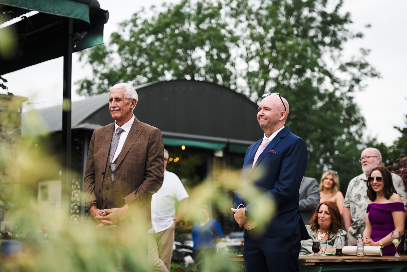 A Relaxed Summer Wedding at Village Barn, Tyrellspass | Amy & Eoghan 101 Two older men in suits stand outdoors at an event, with several seated and standing guests in the background. Greenery and a building are also visible.