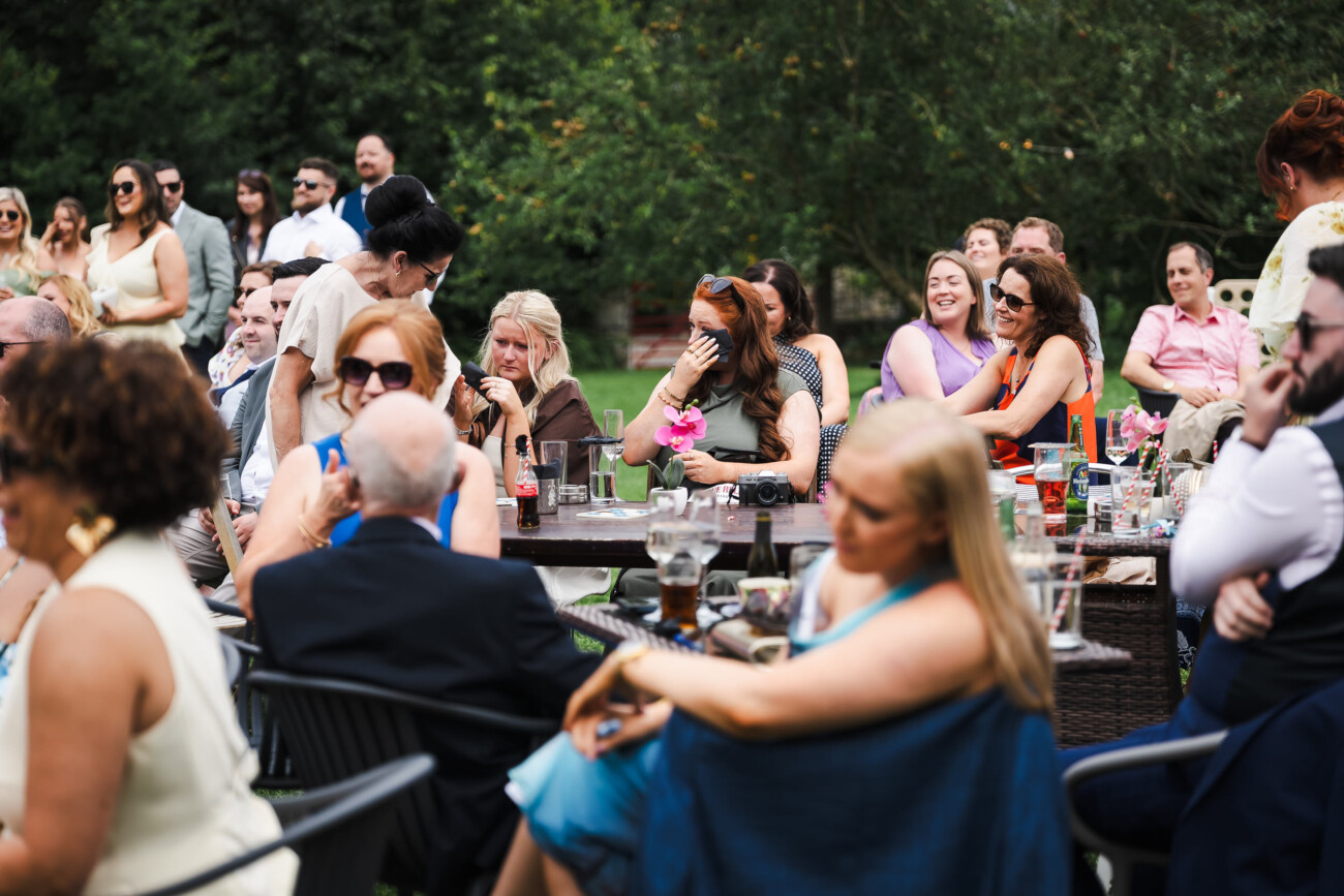 A Relaxed Summer Wedding at Village Barn, Tyrellspass | Amy & Eoghan 102 People sitting outdoors around tables at a gathering, some talking, some watching something off-camera, with drinks and greenery in the background.