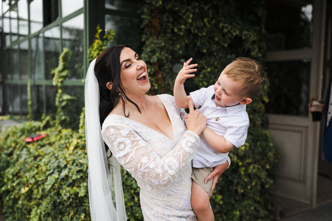 A Relaxed Summer Wedding at Village Barn, Tyrellspass | Amy & Eoghan 103 A woman in a white dress holds a laughing young boy outside near greenery, both smiling and interacting playfully.
