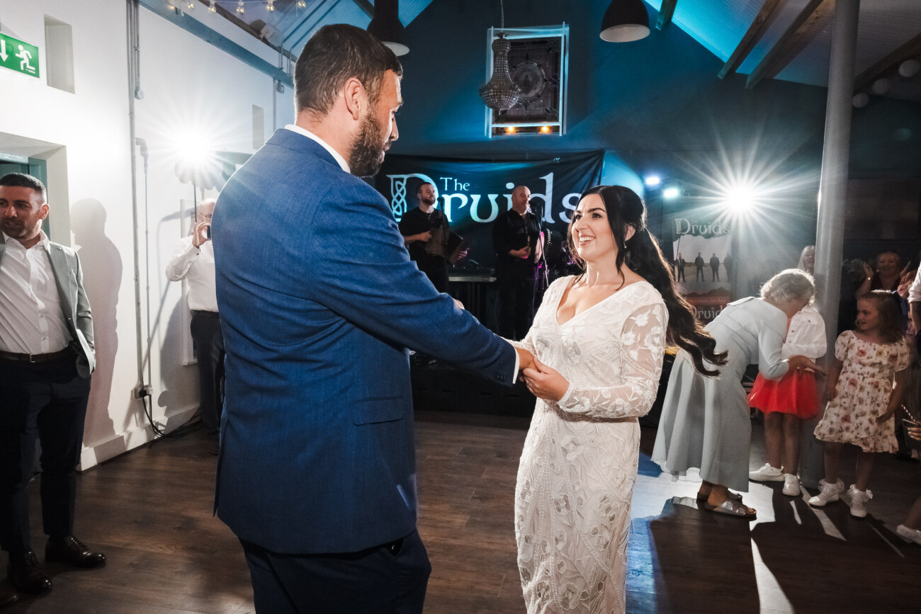 A Relaxed Summer Wedding at Village Barn, Tyrellspass | Amy & Eoghan 108 A bride and groom dance together in a well-lit room, surrounded by guests watching and smiling.