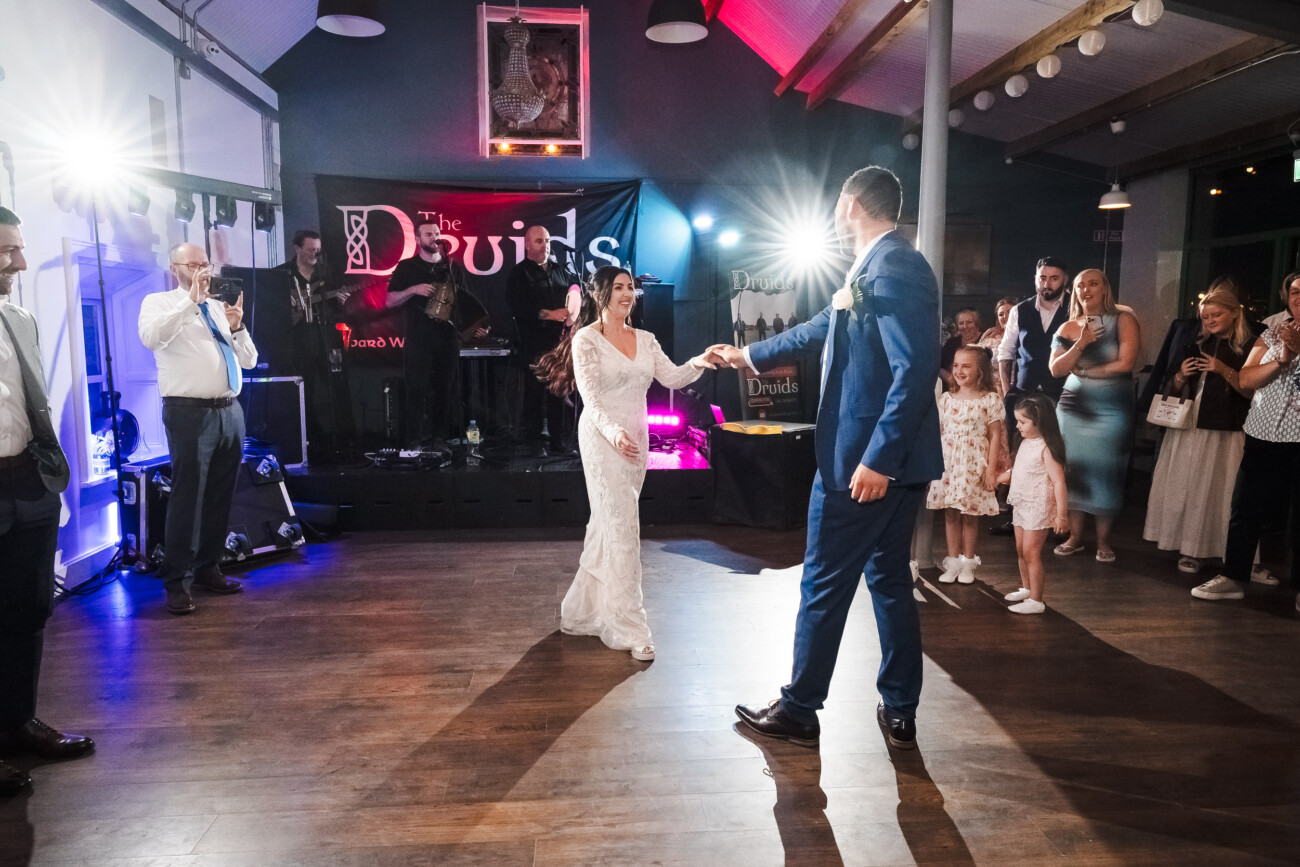 A Relaxed Summer Wedding at Village Barn, Tyrellspass | Amy & Eoghan 110 A bride and groom dance in the centre of a room, surrounded by guests and a live band. Bright lights illuminate the scene.