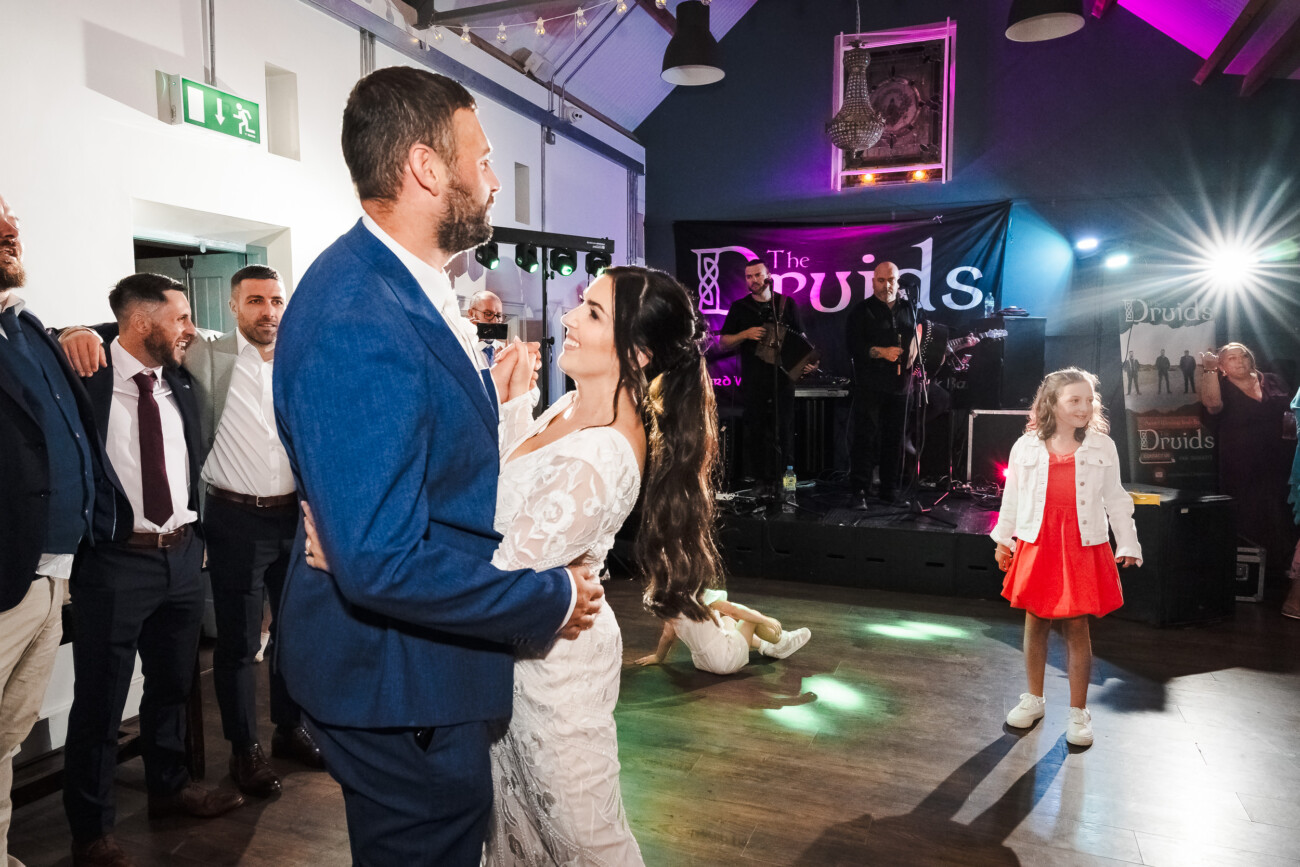 A Relaxed Summer Wedding at Village Barn, Tyrellspass | Amy & Eoghan 111 Bride and groom dance together at a wedding reception while guests and a band called The Druids watch; a young girl in a red dress stands nearby.